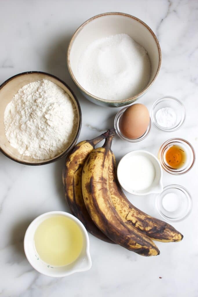 ingredienten voor een bananencake: drie overrijpe bananen en schaaltjes met ingredienten als bloem, suiker, ei, olie, bakpoeder en vanille extract