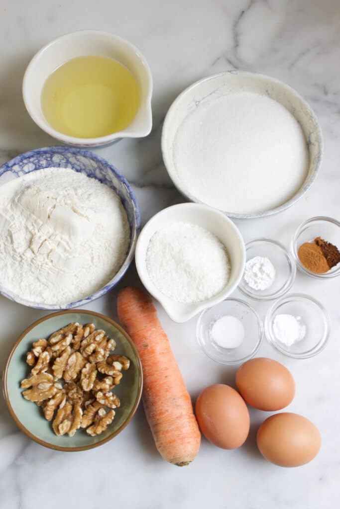 Kommetjes met ingredienten voor een worteltaart: olie, suiker, bloem, kokos, zout, bakingsoda, bakpoeder, walnoten, kaneel en kruidnagel. Drie losse eieren en een winterwortel.