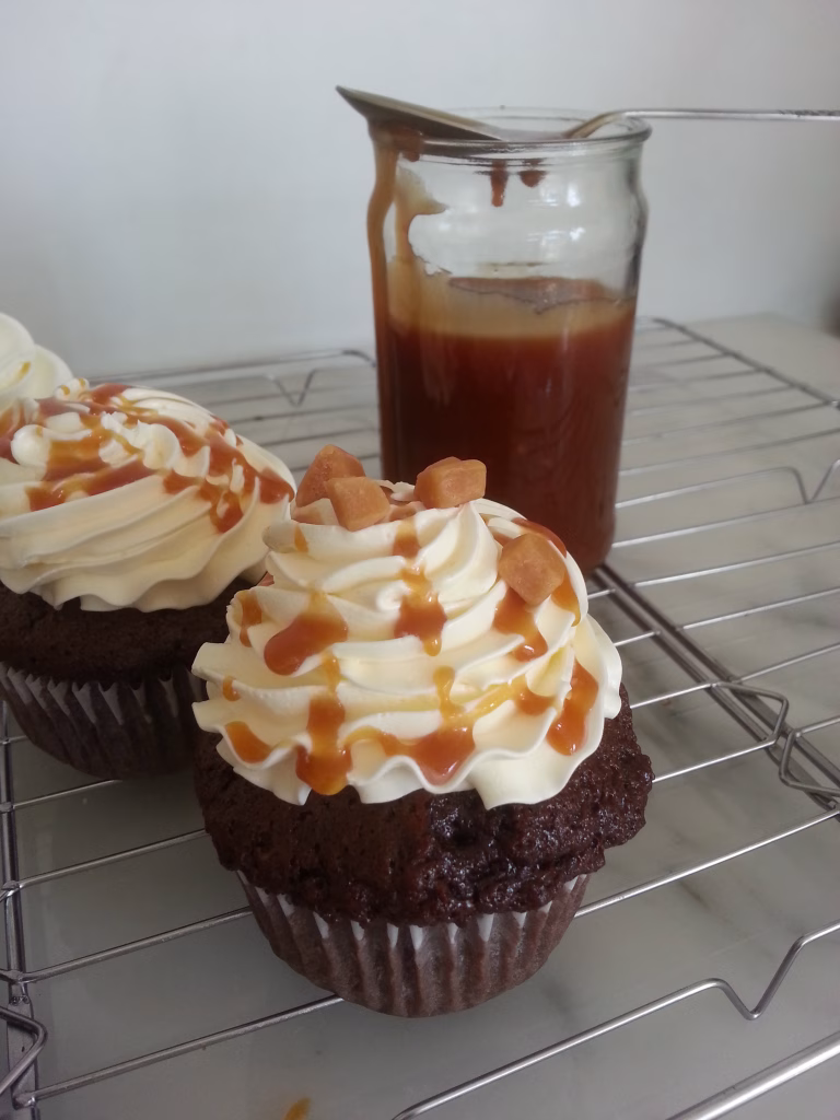 frappuccino cupcakes gedecoreerd met een toef botercrime, karamelsaus en karamel blokjes op een taartrooster. Er achter staat een potje met karamelsaus.