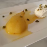 gepocheerde peren van ottolenghi zijn mooi geel door de saffraan. Geserveerd op een wit bord met pistachenoten en slagroom.