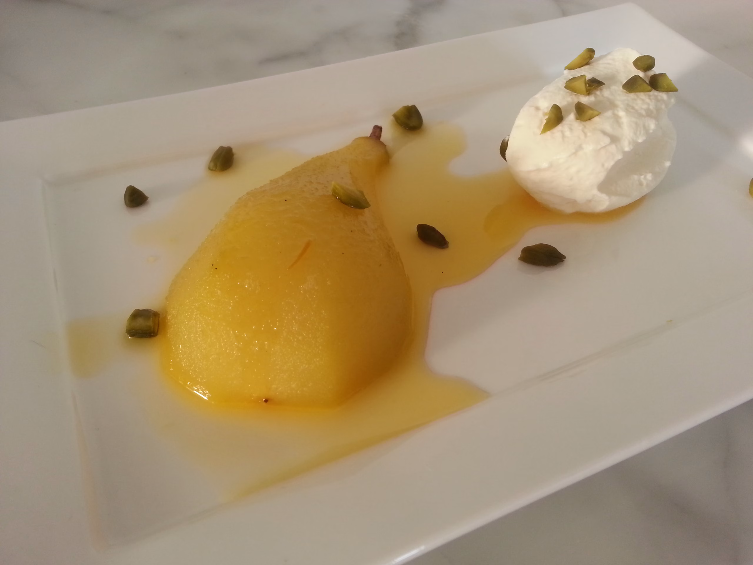 gepocheerde peren van ottolenghi zijn mooi geel door de saffraan. Geserveerd op een wit bord met pistachenoten en slagroom.