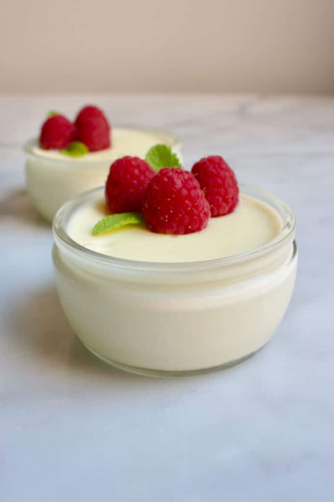 Twee glazen schaaltjes met limoencreme erin gedecoreerd met elk drie frambozen en een takje munt. 