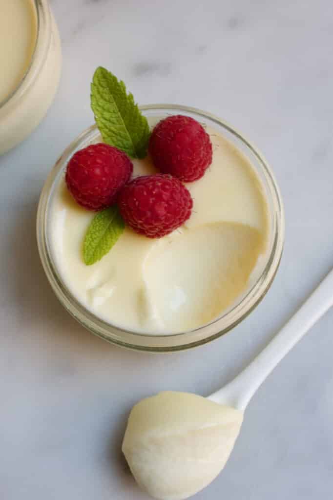 Een schaaltje met limoencreme erin gedecoreerd met drie frambozen en een takje munt. Voor het schaaltje ligt een lepel met deze lemon posset erop