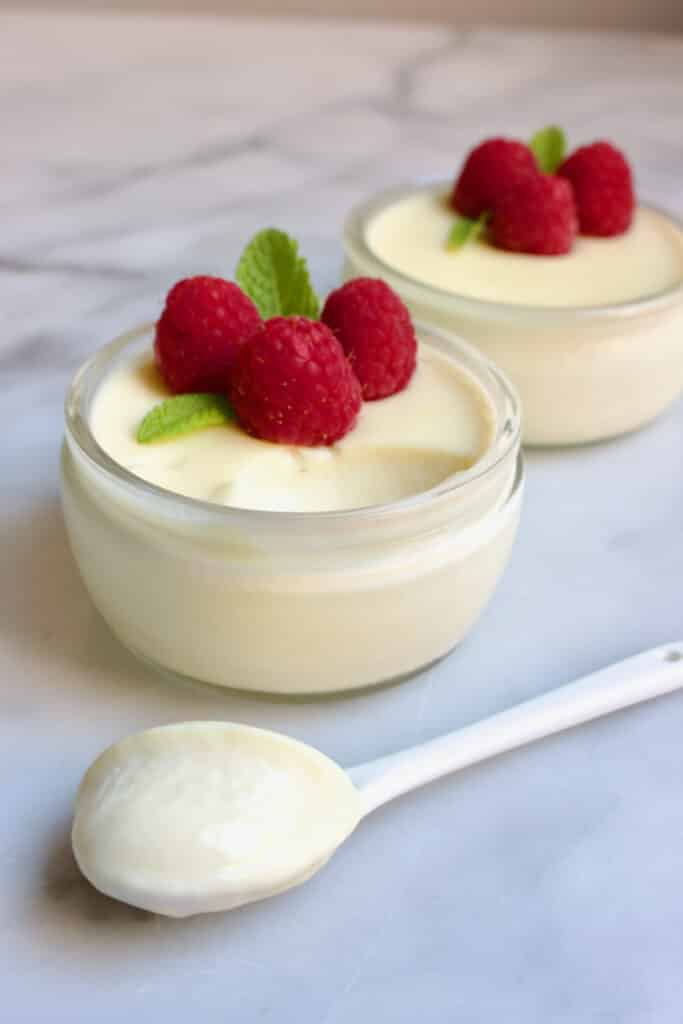 Twee glazen schaaltjes met limoencreme erin gedecoreerd met elk drie frambozen en een takje munt. Voor de schaaltjes ligt een lepel met deze lemon posset erop