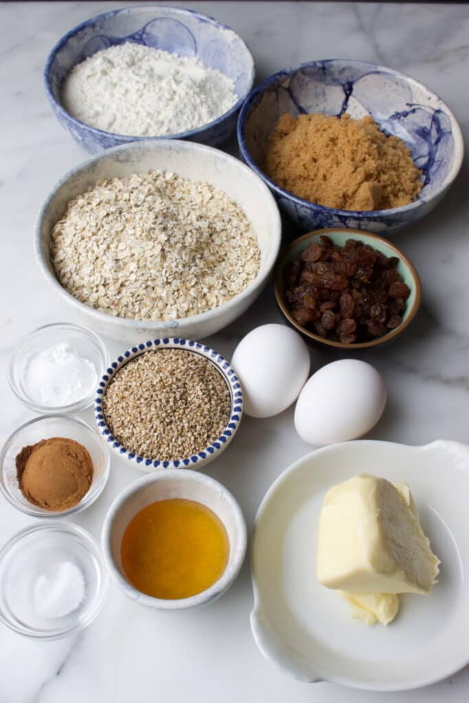 bakjes met ingredienten om zachte havermout koeken te maken: bruine basterdsuiker, bloem, havermout, rozijnen, sesamzaad, eieren, bakpoeder, zout, koekkruiden, honing en boter