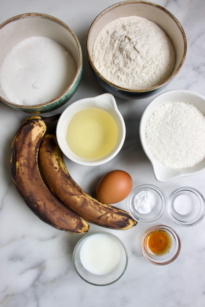 Ingredienten voor een kokos bananencake: rijpe bananen, bakjes met bloem, suiker, kokos, een los ei, glazen schaaltjes met bakingsoda, zout, vanille extract en karnemelk