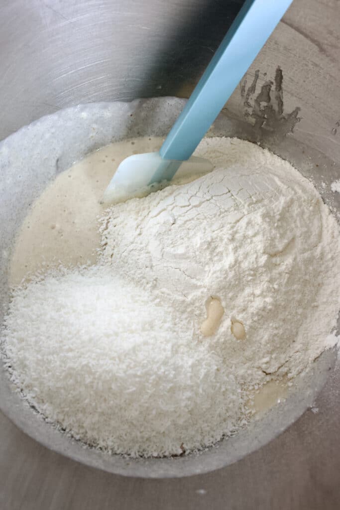 beslag voor een bananencake met bloem en kokos erop om er doorheen te spatelen met een blauwe spatel