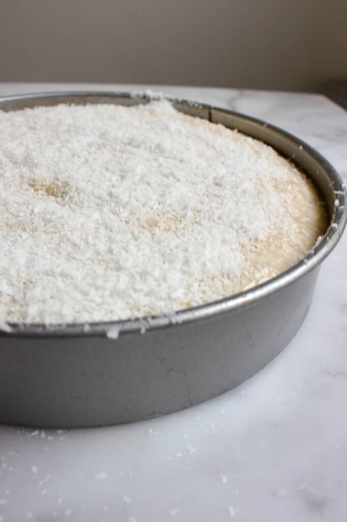 ronde bakvorm met het beslag voor een kokos bananencake erin, wat bestrooid is met extra kokosrasp.
