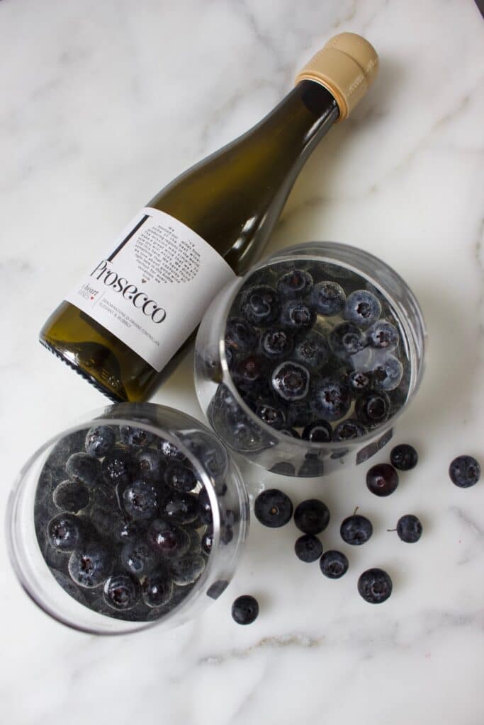 flesje prosecco naast twee glazen die gevuld zijn met blauwe bessen in wijn gelei
