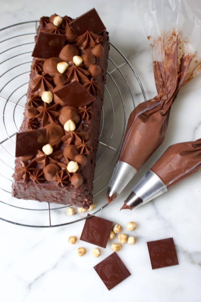 een chocolade cake met een Ferrero Rocher chocolade glazuur en chocolade creme bovenop, twee spuitzakken met de creme liggen er naast en wat losse chocolade decoraties en hazelnoten