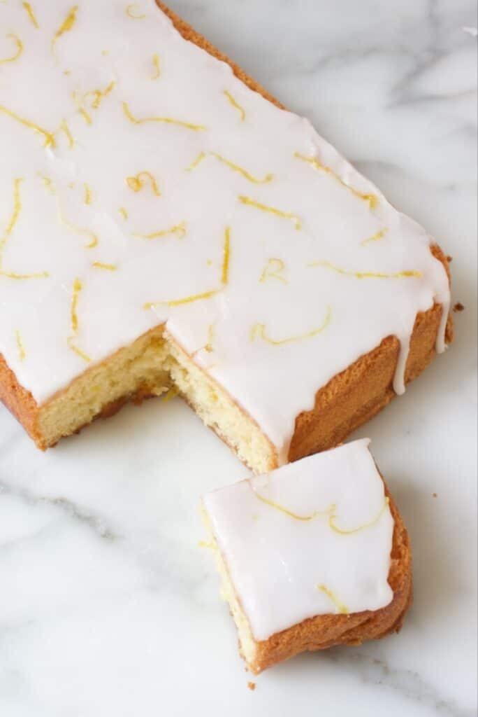 lemon drizzle cake op een marmeren plaat waar een vierkant hoekje uit gesneden is