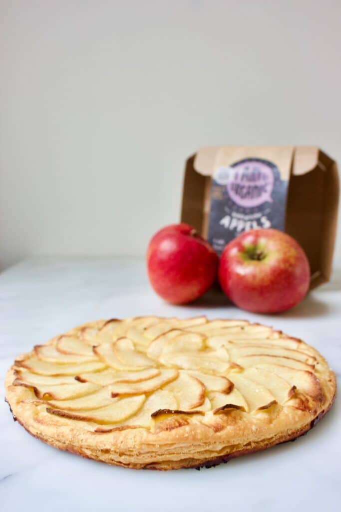 gebakken snelle appeltaart met een bladerdeegbodem en dunne plakjes appel erop in waaiervorm en twee losse appels met een kartonnen appelverpakking erachter