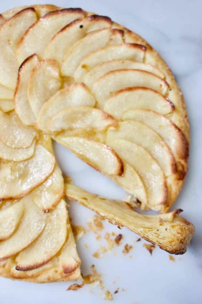 Gebakken ronde tarte fine aux pommes waar een taartpuntje is uitgesneden. Een snelle appeltaart op een bladerdeeg bodem met daarom in waaiervorm dunne schijfjes appel,