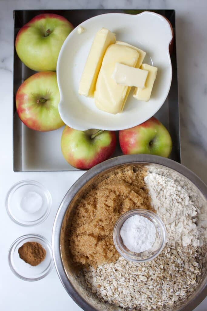 ingredienten voor een appeltaart met havermout: vierkante bakvorm met appels en een schaaltje boter. Ronde kom met suiker, bloem, havermout en een bakje met bakpoeder.