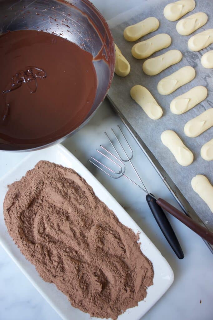 vulling voor slagroomtruffels op een metalen plaat, een kom gesmolten chocolade, een bord met cacaopoeder en twee bonbonvorkjes