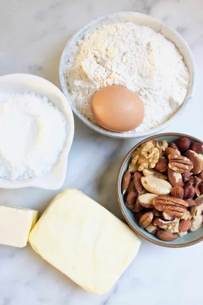 ingredienten voor een boterkoek met noten: pakje boter, schaaltje met gemengde noten, een schaaltje met bloem en een ei en een bakje met witte basterdsuiker
