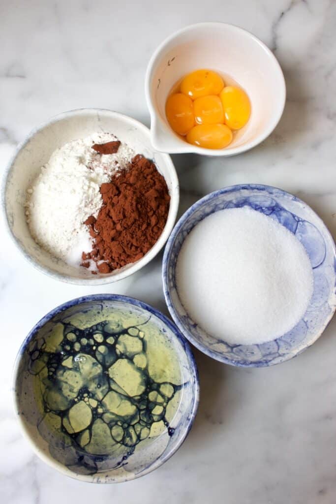 Ingredienten voor chocolade biscuit in schaaltjes: eidooier, suiker, bloem en cacao, eiwit
