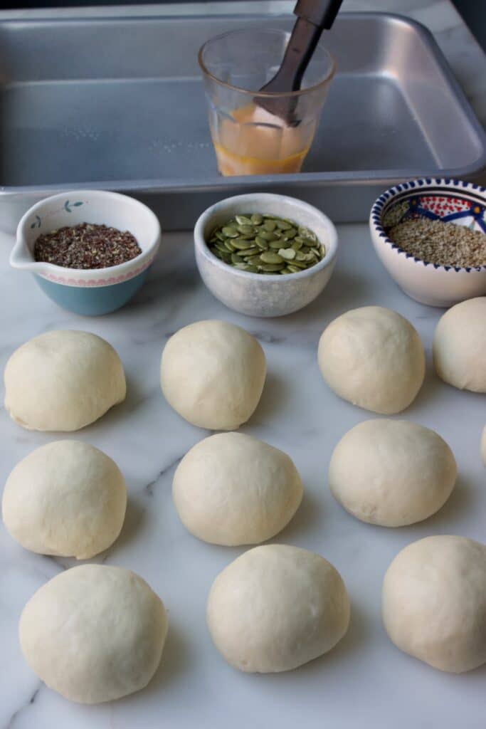 tien deegbolletjes van wit brooddeeg  met kleine bakjes gevuld met lijnzaad, pompoenpitten en sesamzaad. Daarachter een glas met losgeklopt eiwit en een kwastje erin zodat de broodjes bestreken kunnen worden met ei en daarna in de pitten/zaden gedoopt kunnen worden.