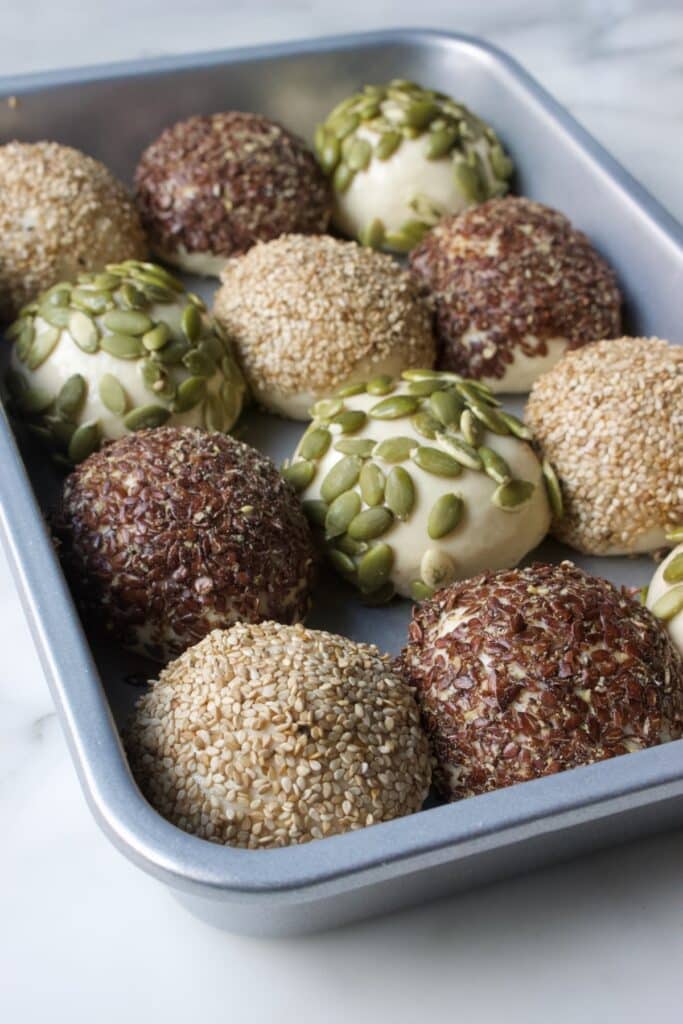 een bakvorm met bolletjes breekbrood die bedekt zijn met pompoenzaad, sesamzaad of lijnzaad, klaar om te rijzen.