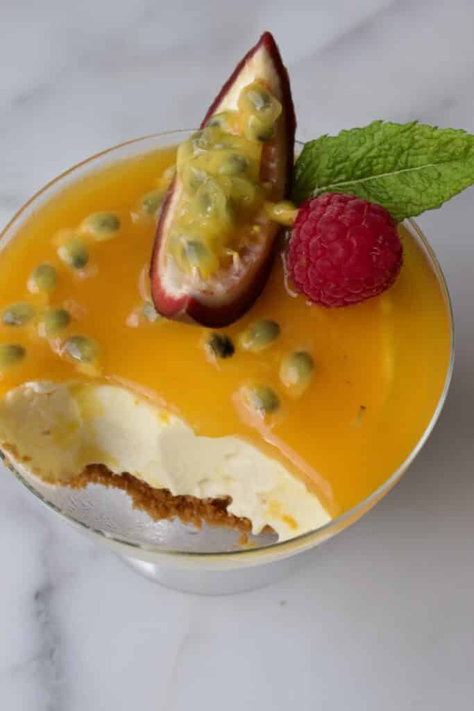 glaasje met cheesecake met passievrucht toetjes gedecoreerd met passievrucht, munt en framboos waarbij er al een beetje uitgelepeld is.