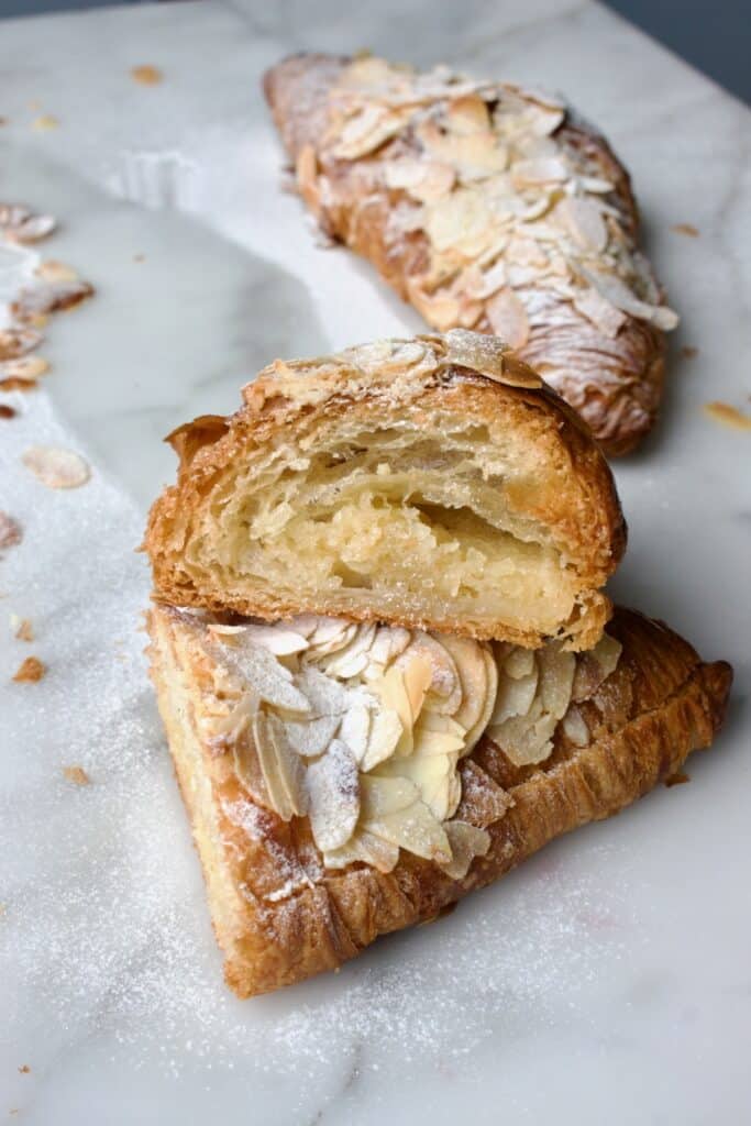 doorgesneden amandel croissant zodat je de amandelvulling kunt zien. op de achtergrond een hele amandel croissant