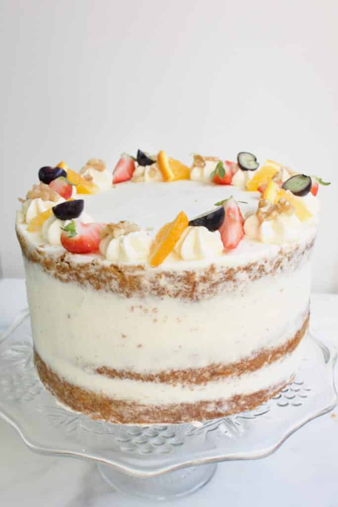 een carrot cake taart met drie lagen cake en roomkaas creme ertussen en gedecoreerd met fruit en noten. Op een glazen taart plateau.