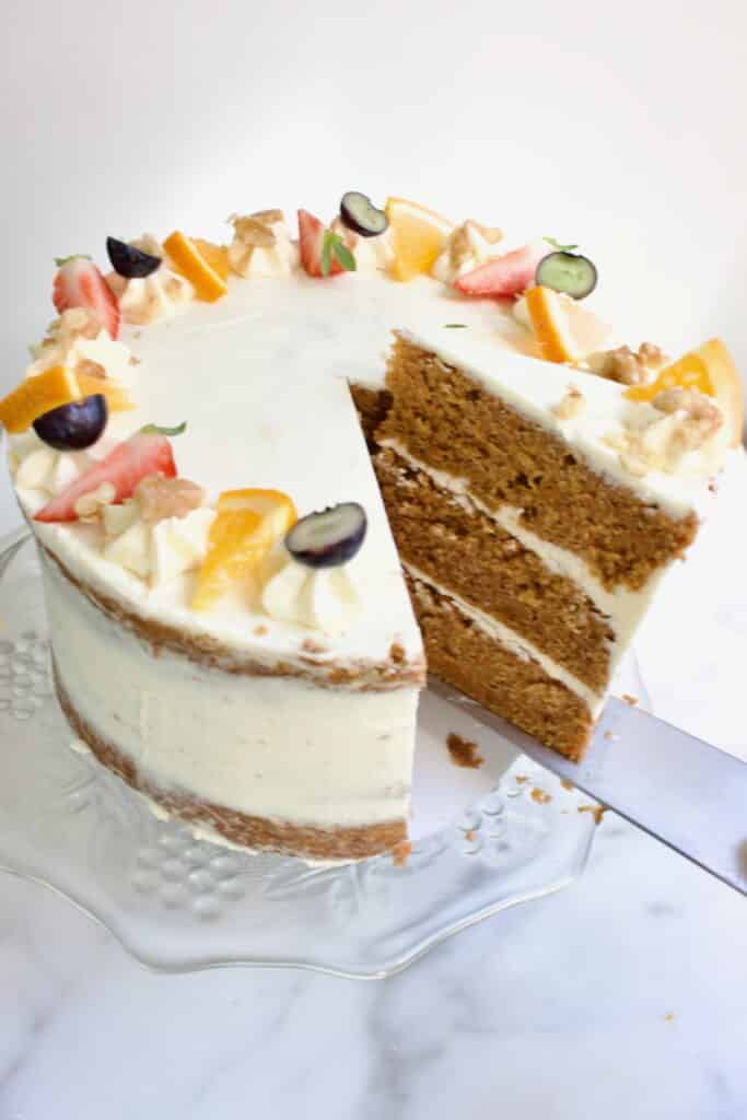 een carrot cake taart met drie lagen cake en roomkaas creme ertussen en gedecoreerd met fruit en noten, waarbij een taartpunt uit de taart gehaald wordt