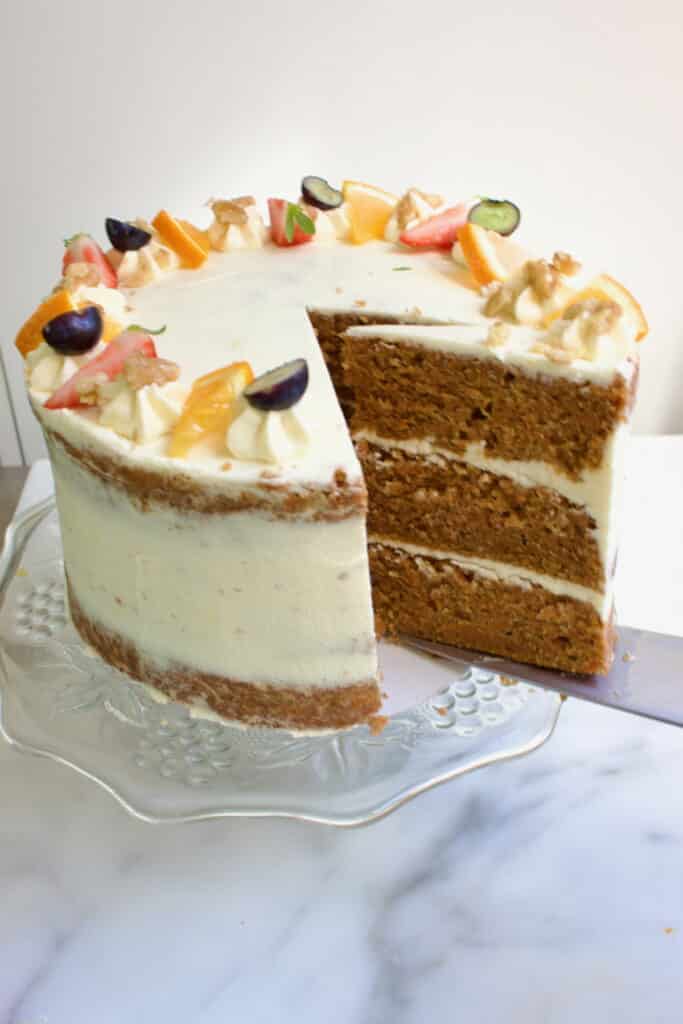 een carrot cake taart met drie lagen cake en roomkaas creme ertussen en gedecoreerd met fruit en noten, waarbij een taartpunt uit de taart gehaald wordt