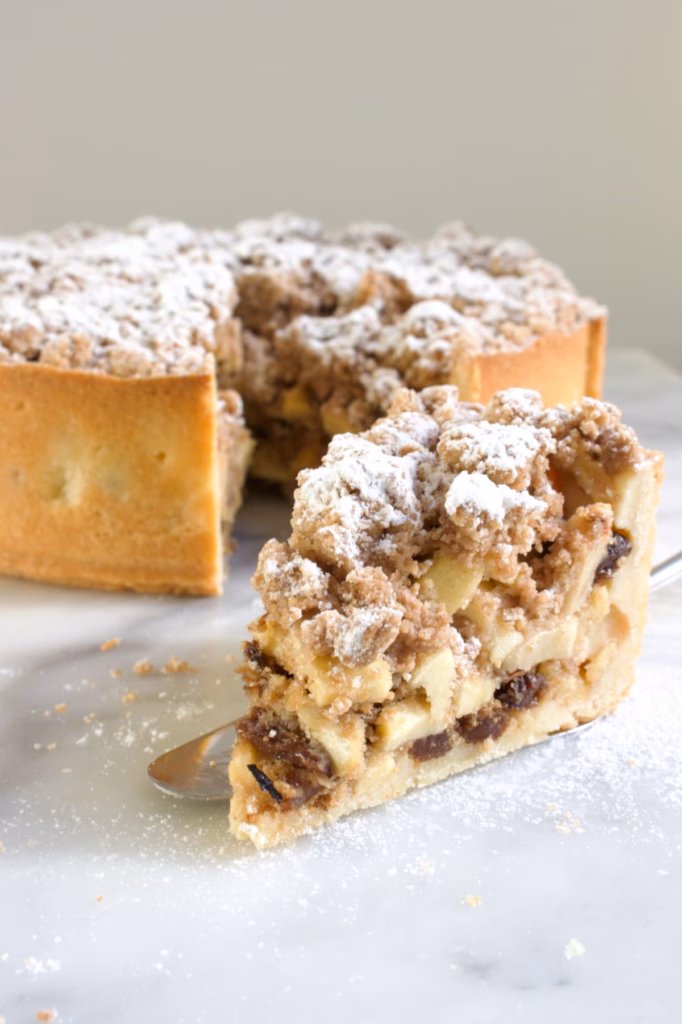 Een punt appeltaart met rozijnen en een kruimellaag erop met speculaaskruiden en poedersuiker. Op de achtergrond staat de hele taart.