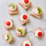 stukjes traditionele Friese oranjekoek. Vierkant, met roze glazuur en gedecoreerd met slagroom en een framboos of stukje kiwi.